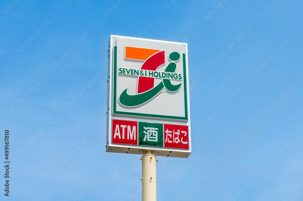 日本のセブン＆アイ・ホールディングスコンビニエンスストア看板の詳細写真。上部には「SEVEN & i HOLDINGS」のロゴとテキスト、下部の赤いパネルには大きな赤と白の文字でサービスリスト「ATM」、それに続いてサービスアイコン（カードを持つ手）、そして単語「酒」（日本酒・アルコール）とボトル/カップアイコン、最後に「たばこ」とタバコのアイコンが記載されています。